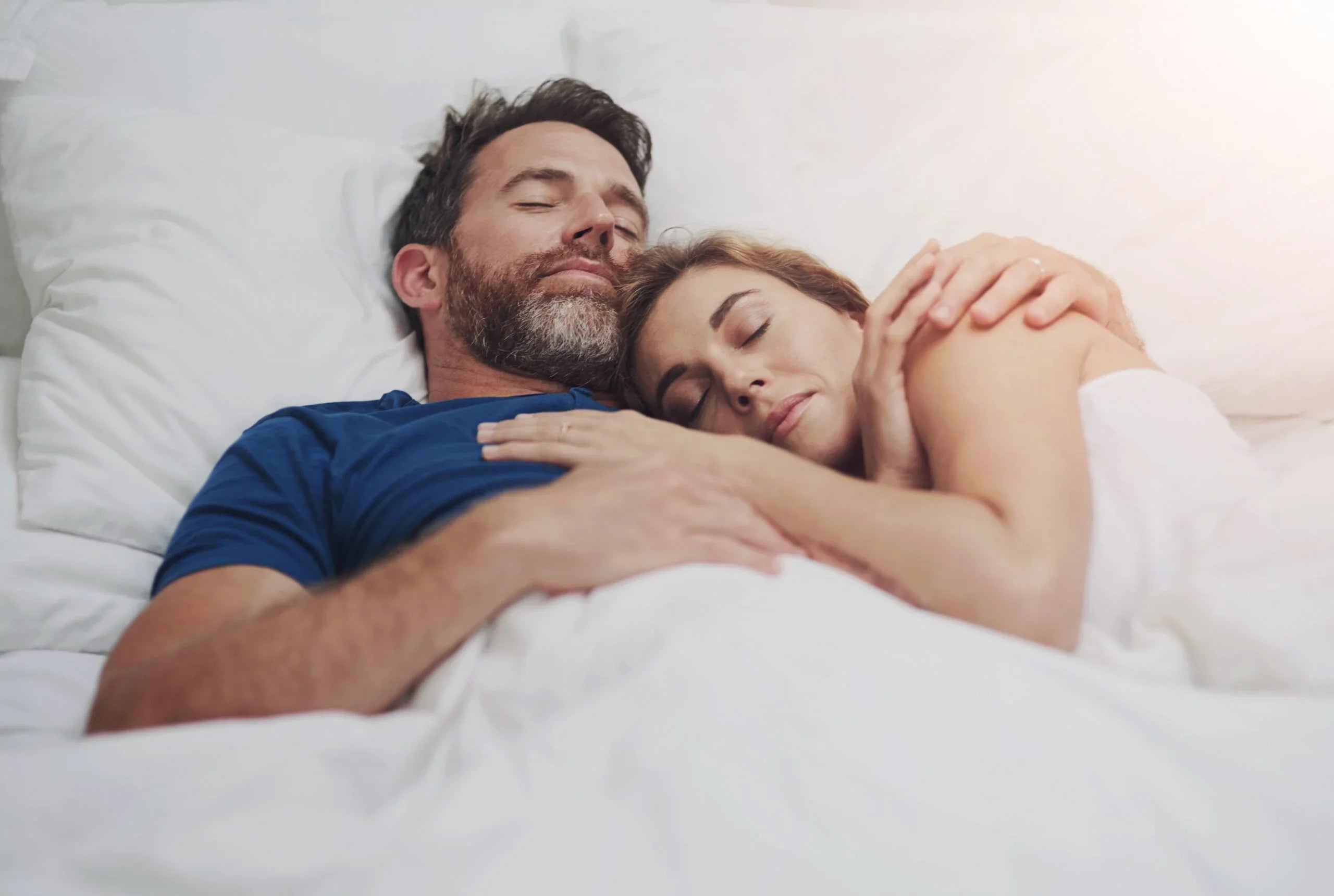 Couple lying in bed cuddling with eyes closed, relaxed and comfortable under white sheets