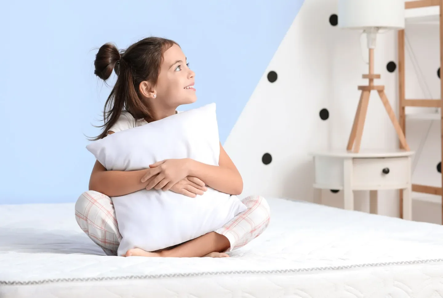 Smiling girl holding white pillow sitting cross-legged on bed in modern bedroom with polka dot wall
