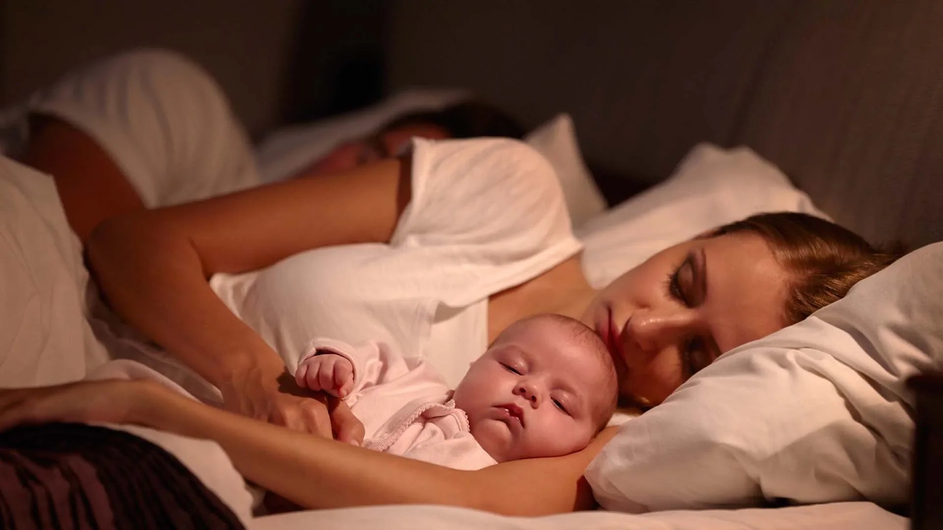 Mother and father sleeping in bed holding their baby peacefully under soft lighting