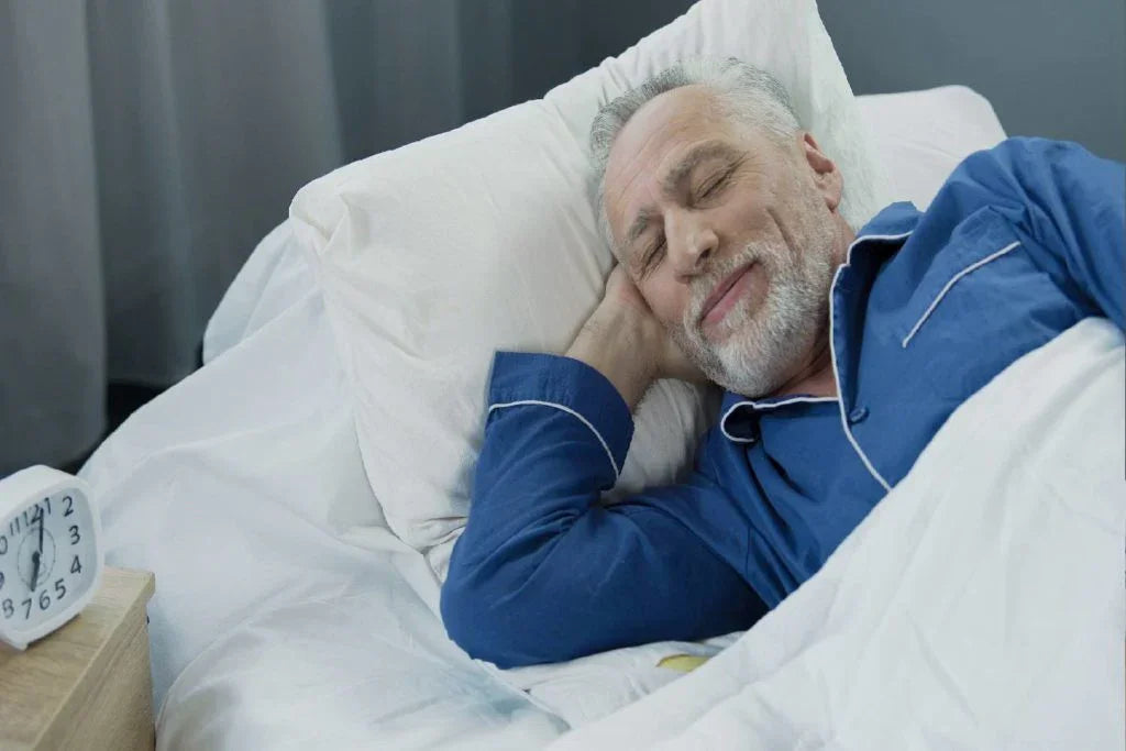 Elderly man in blue pajamas sleeping peacefully in white bed with alarm clock on nightstand