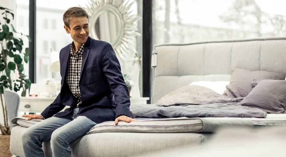 Smiling man sitting on a gray mattress in a bright mattress showroom with modern decor