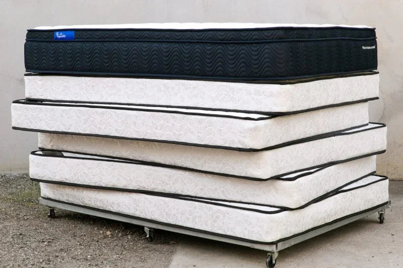 Stack of five mattresses with white patterned covers and one black mattress on top, displayed outdoors