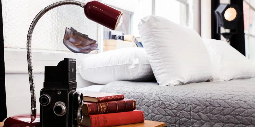 Modern bedroom with grey quilted bedspread, white pillows, vintage camera, red lamp, and stacked books