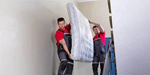 Two men in red shirts carrying a mattress downstairs in a white-walled interior