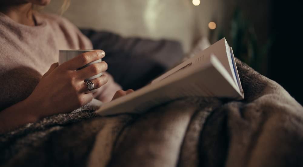 Woman relaxing with a cup of tea and reading a book under a soft blanket in cozy home setting