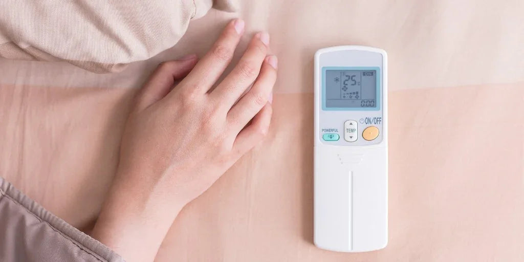 Woman's hand next to white air conditioner remote control set at 25 degrees on beige bed