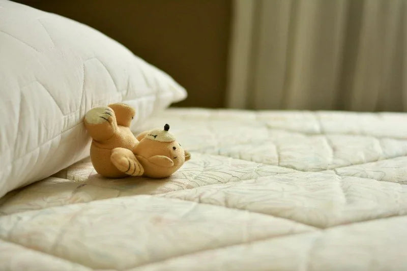 Small plush teddy bear lying on a quilted mattress next to a white pillow in a soft-lit bedroom