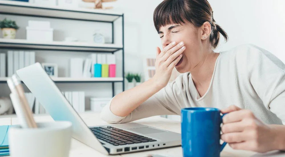 Tired woman yawning while holding blue mug at desk with laptop illustrating sleep deprivation effects