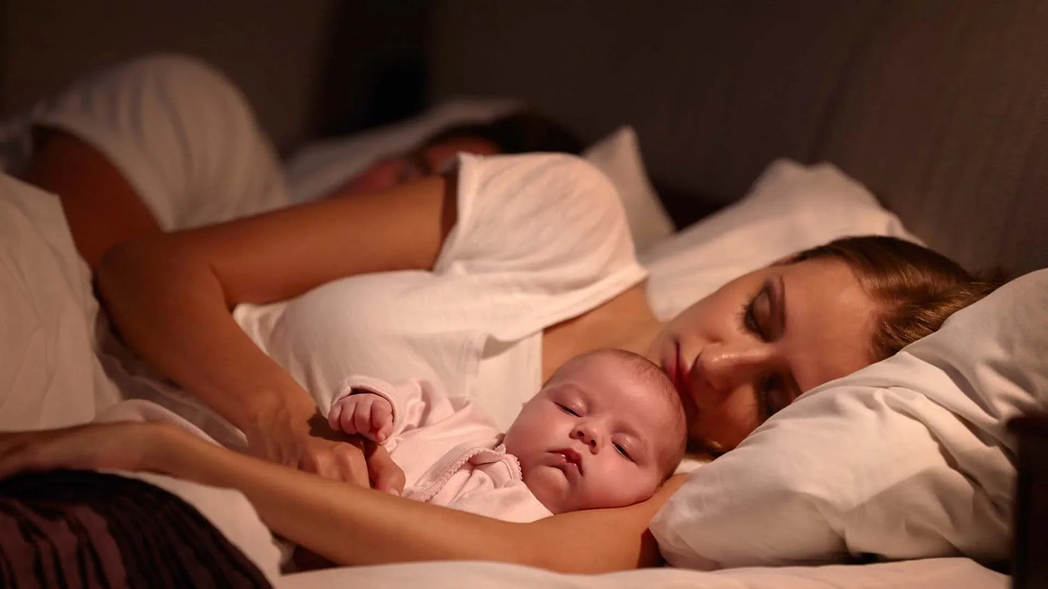 Mother and father sleeping in bed holding their baby peacefully under soft lighting