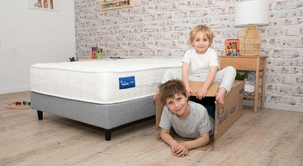 Two children playing with a cardboard box near a white mattress bed in a bright, modern bedroom