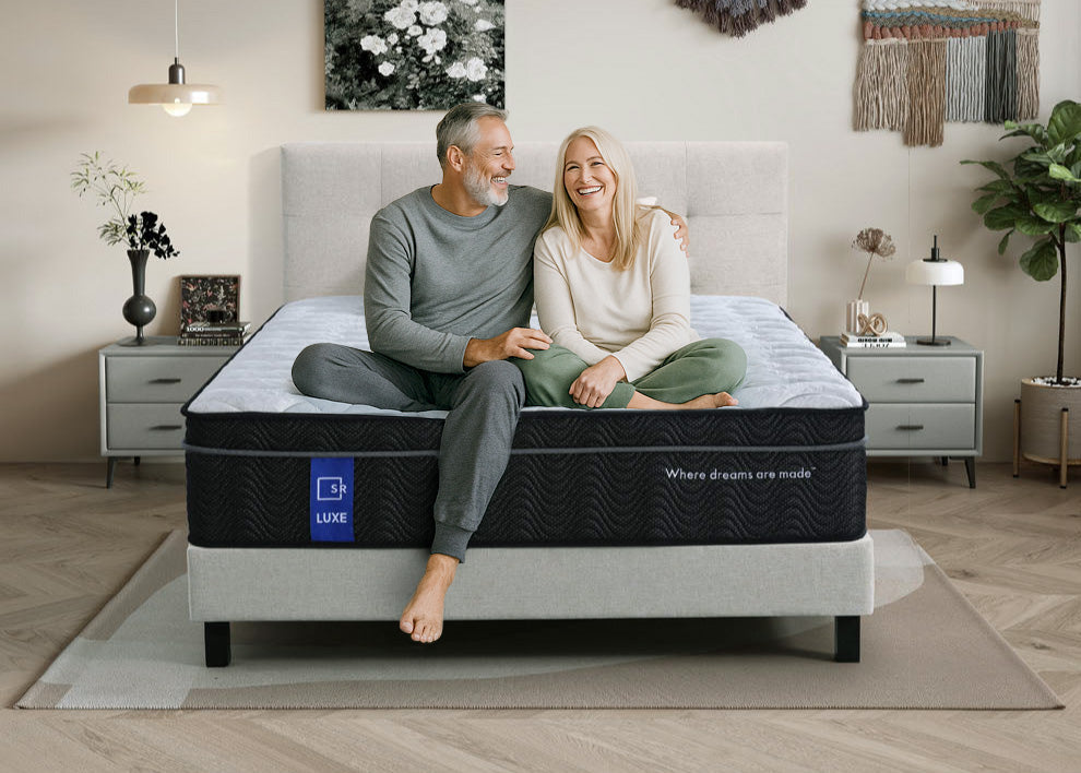 Smiling middle-aged couple sitting on SR Luxe mattress in modern bedroom with plants and nightstands