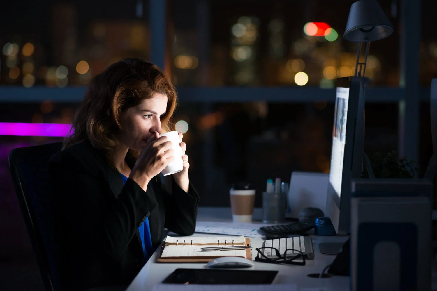 Woman drinking coffee while working late at office desk with computer and notebook