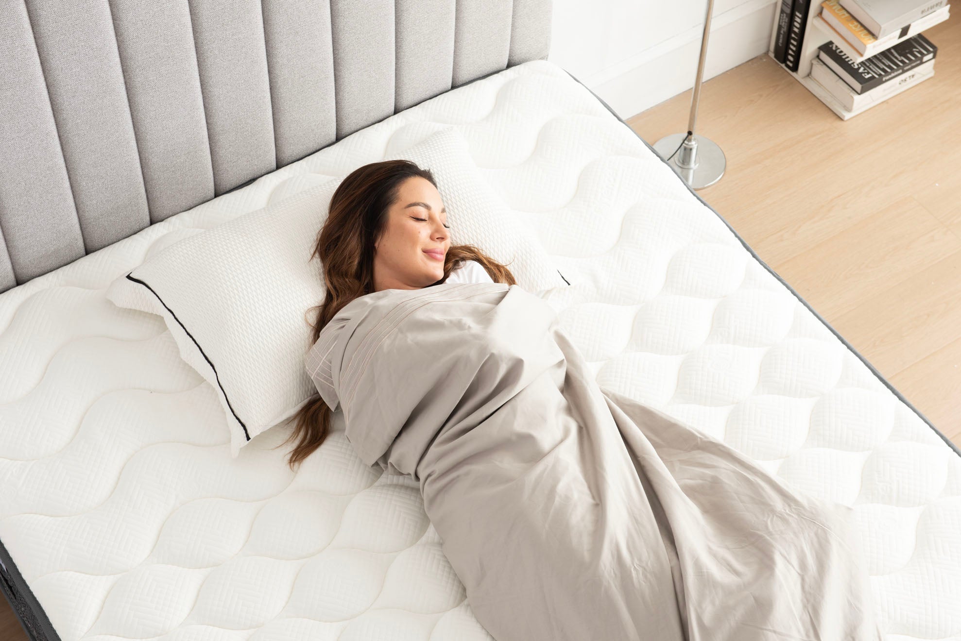 Woman sleeping wrapped in grey blanket on white quilted mattress with grey tufted headboard