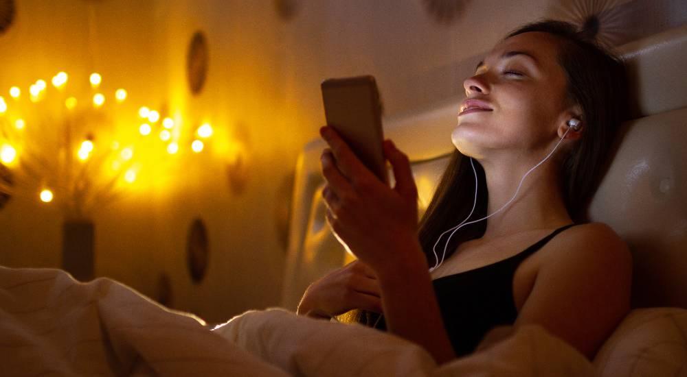 Woman relaxing in bed with earphones listening to music on smartphone in warm cozy room lighting