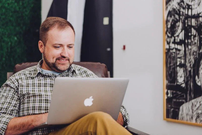 Man with beard using Apple laptop indoors, sitting on chair near abstract black and white painting