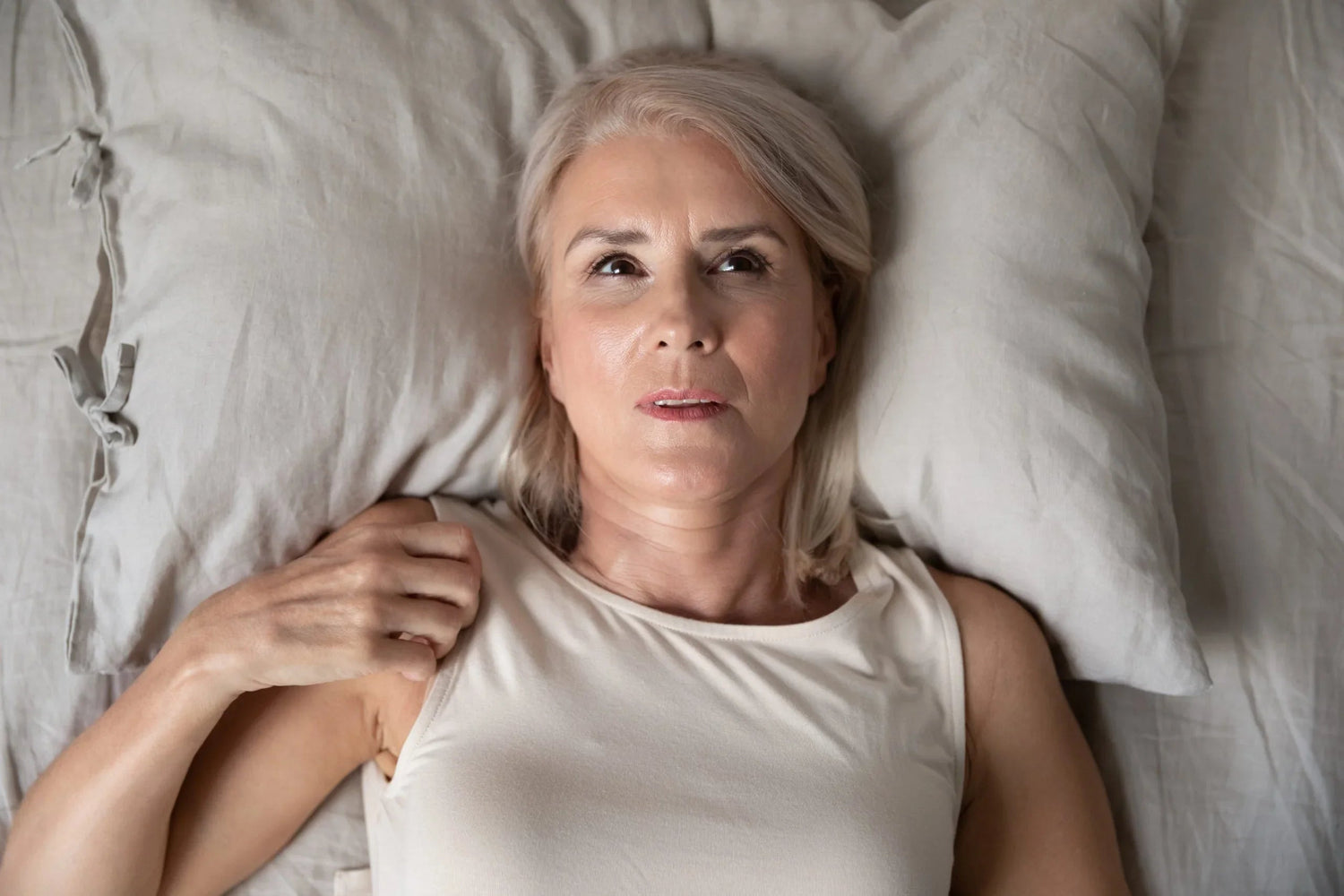 Middle-aged woman lying in bed with a thoughtful expression, wearing a white sleeveless top