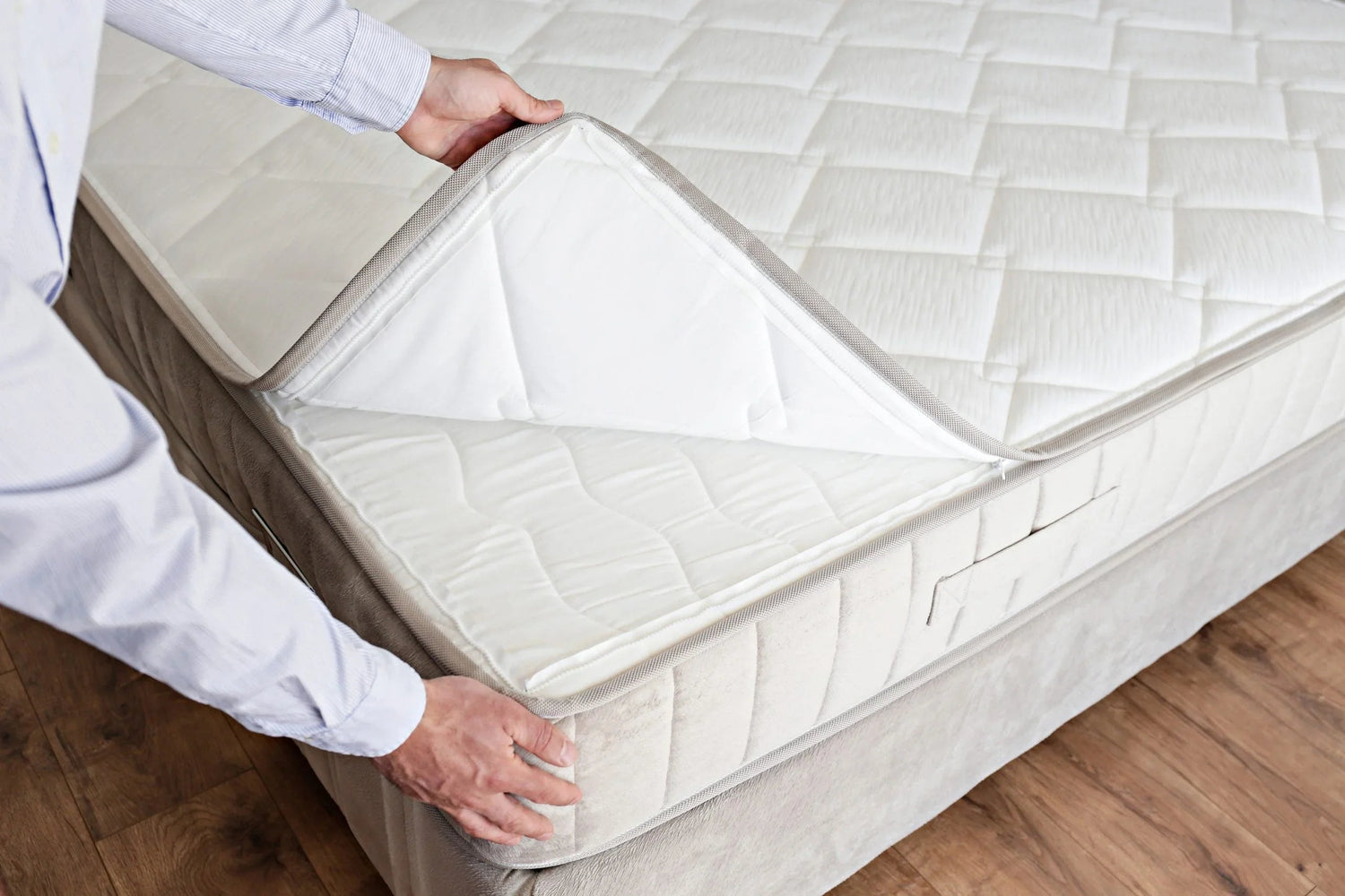 Person lifting quilted mattress topper on beige mattress in bright room with wooden floor