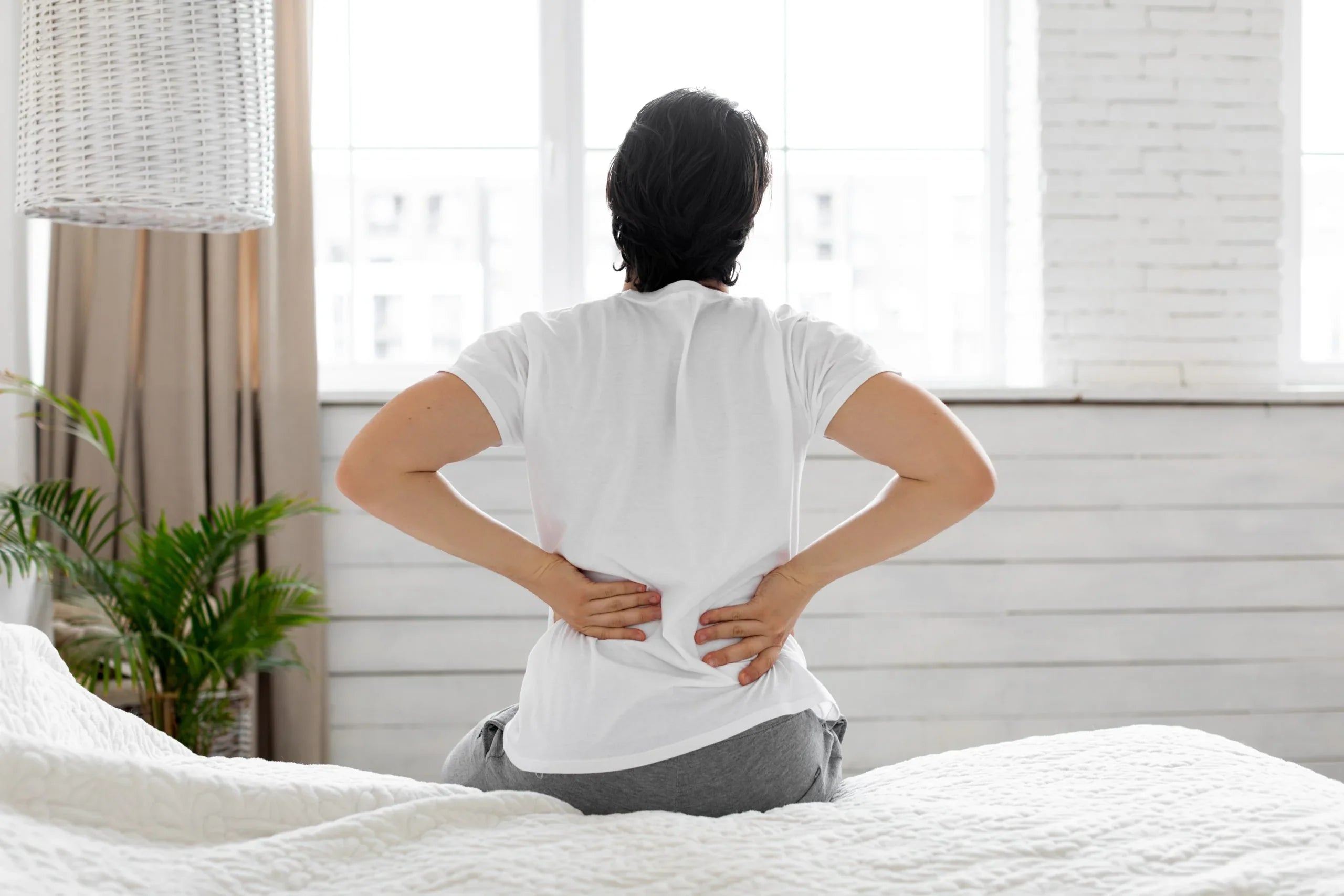Person in white shirt sitting on bed holding lower back in pain in bright bedroom