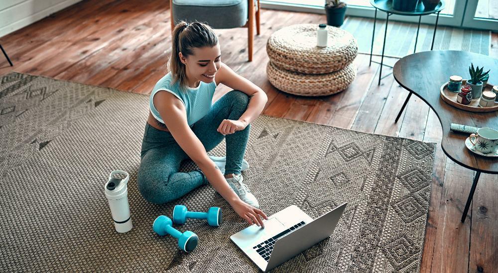 Woman in workout clothes using laptop on carpeted floor with dumbbells and water bottle nearby in cozy living room