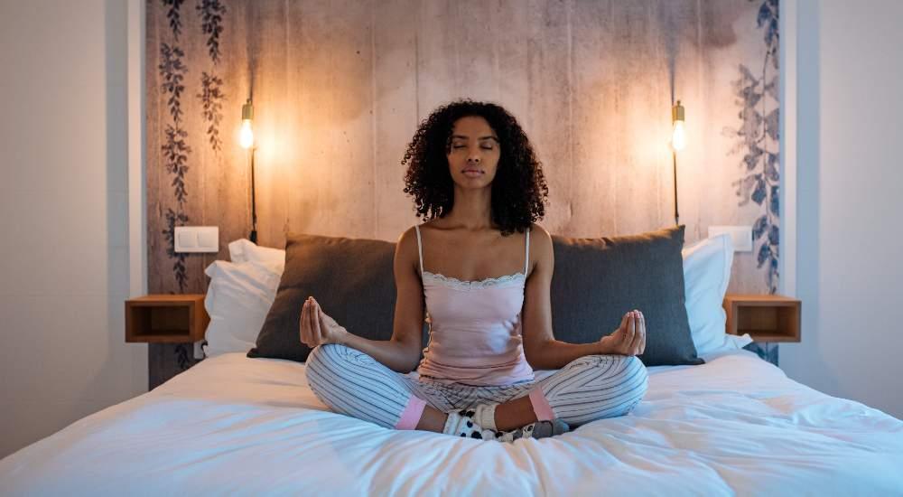 Woman meditating cross-legged on bed in cozy bedroom with warm lighting and neutral decor