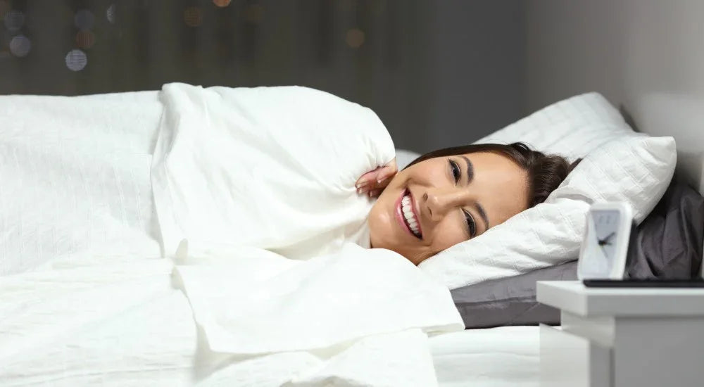 Smiling woman lying in bed wrapped in white blanket with pillow and bedside clock