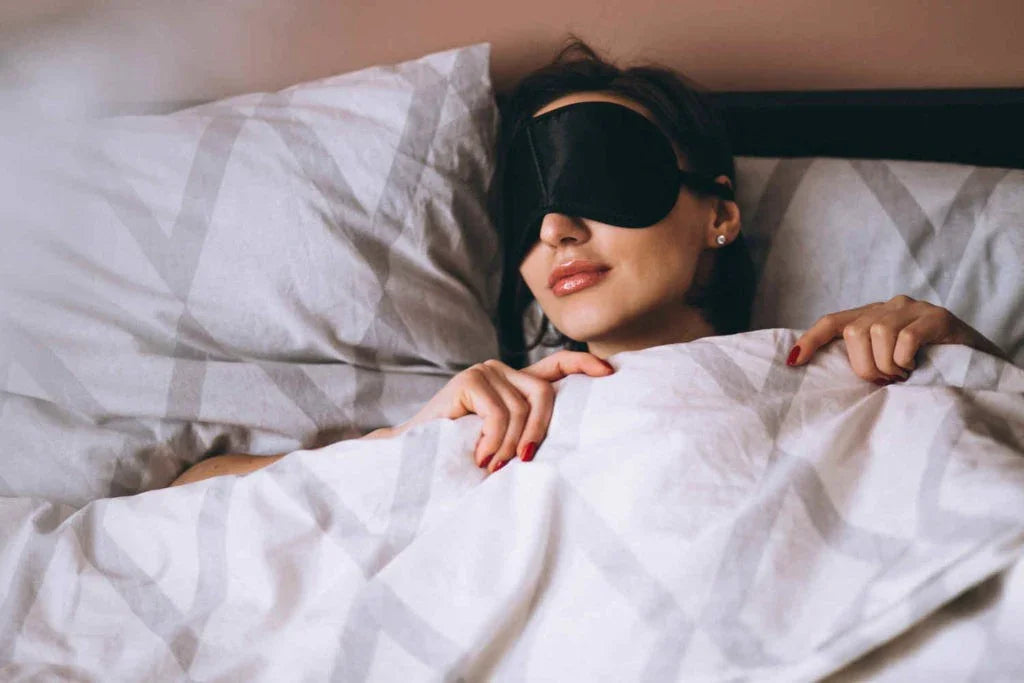 Woman peacefully sleeping in bed wearing a black eye mask under white patterned bedding